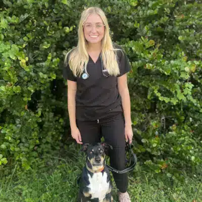 Dr-Marr-profile-400×400.jpg A veterinarian in black scrubs with a stethoscope around her neck stands outside in front of green bushes, smiling and holding the leash of a black and tan dog sitting by her side.