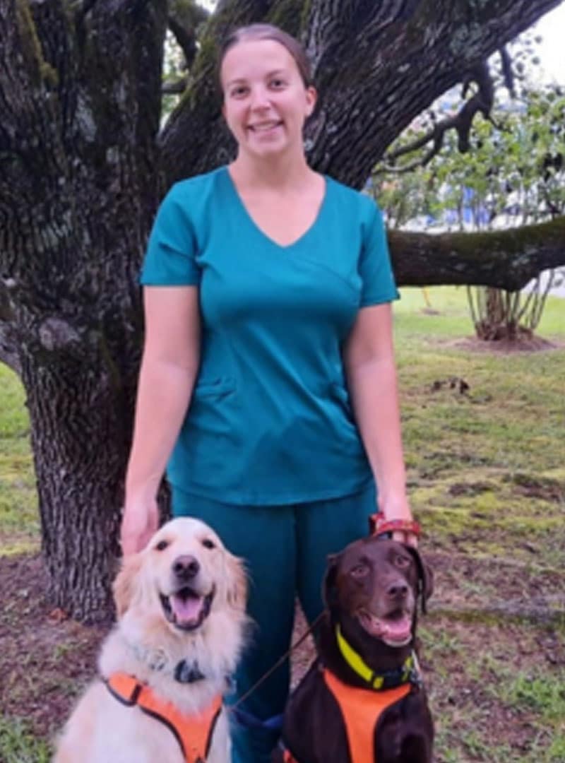 A veterinarian in teal scrubs stands outside in front of a large tree, smiling as she holds two dogs on leashes. One dog is golden with a cream coat, the other is brown; both wear orange harnesses.
