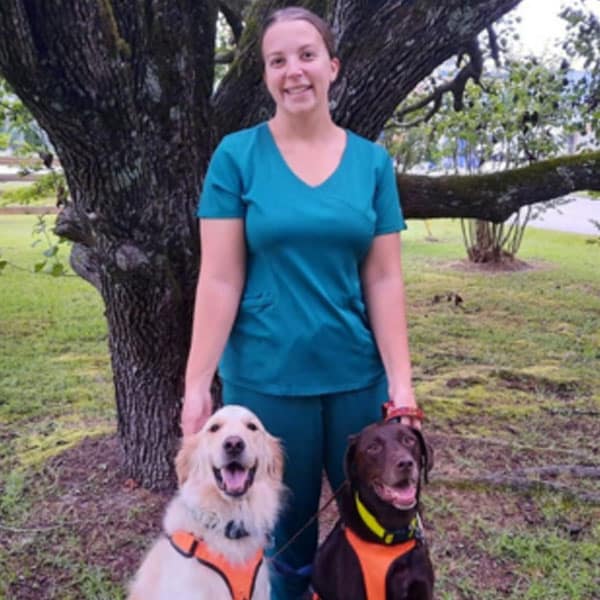 A veterinarian in teal scrubs stands outdoors in front of a tree, smiling while holding two leashed dogs wearing orange harnesses; one dog is golden and the other is brown.