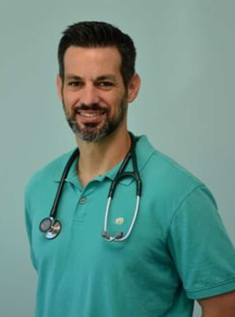 A man with short dark hair and a beard, wearing a teal polo shirt and a stethoscope around his neck, stands smiling in front of a plain light teal background, clearly presenting himself as a friendly veterinarian.