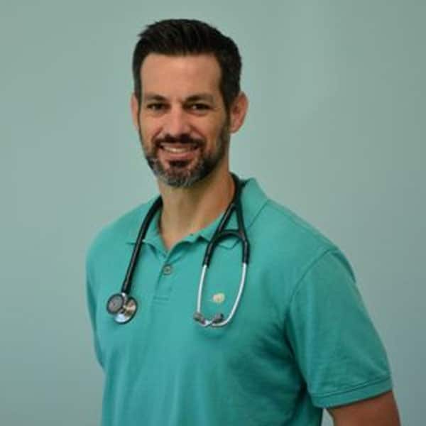 A man with short dark hair and a beard, wearing a teal polo shirt and a stethoscope around his neck, stands smiling against a plain light blue background, embodying the welcoming presence of a dedicated veterinarian.