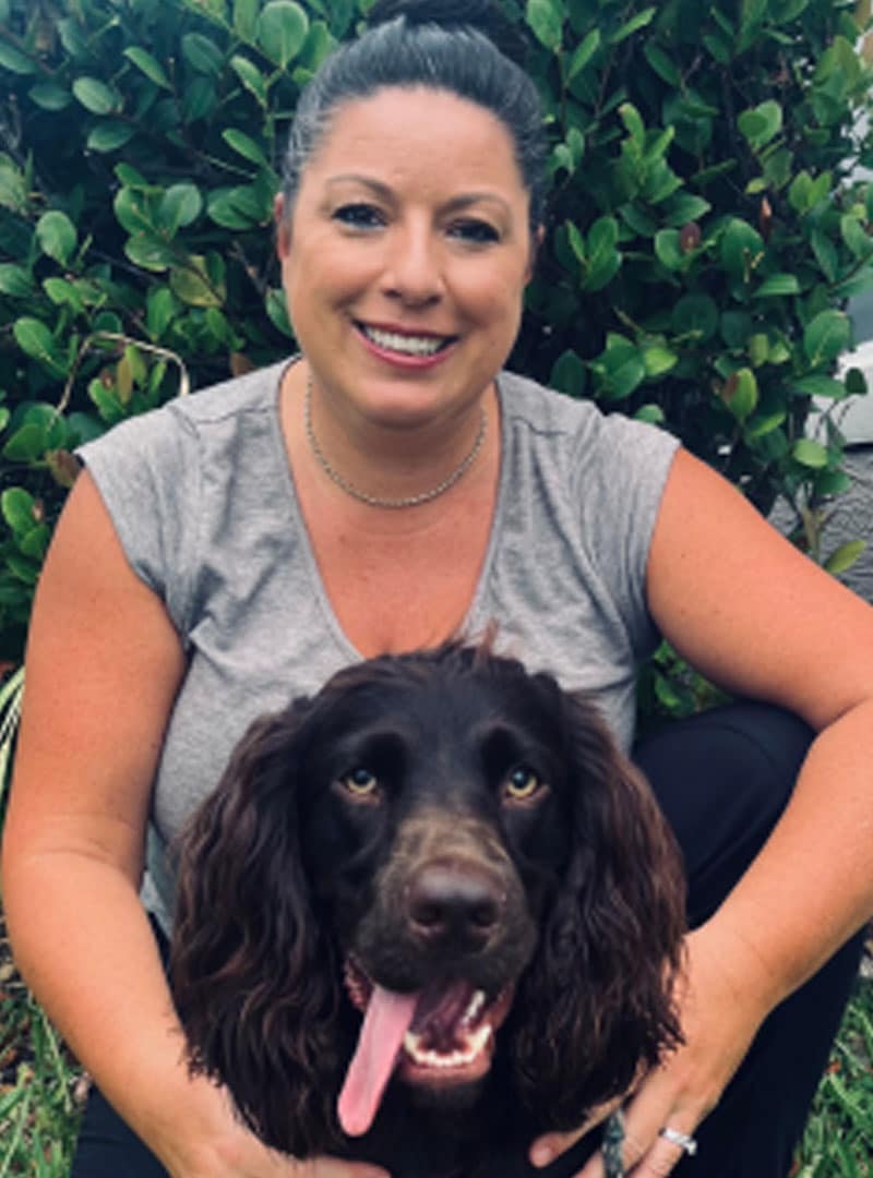 A woman with dark hair in a bun, wearing a gray T-shirt, smiles while kneeling next to a brown dog with long fur and its tongue out. They are outdoors in front of green leafy bushes, looking like a happy veterinarian and her furry friend.
